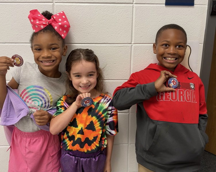 Three children in front of a wall holding small circular objects; one has a Halloween shirt.