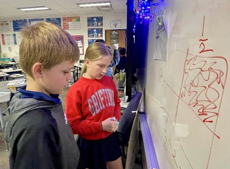Two students look at a whiteboard with drawings, one holding a marker, inside a classroom.