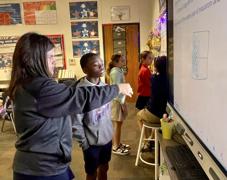 A teacher points to a whiteboard while several children stand nearby and look at the screen.