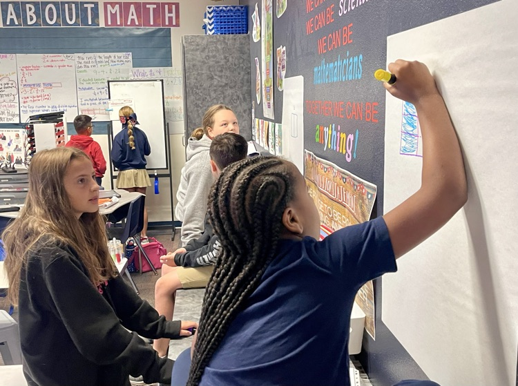 Students in a classroom write on a whiteboard with markers. One student has braids, another is wearing a red hoodie.