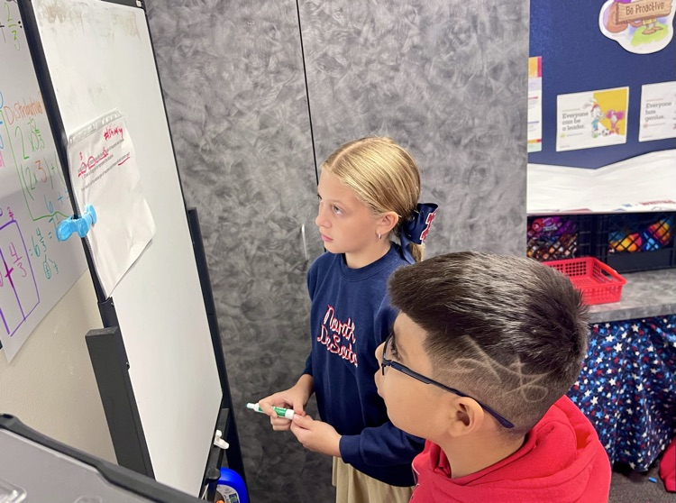 Two children using markers to write on a white board with a blue background.