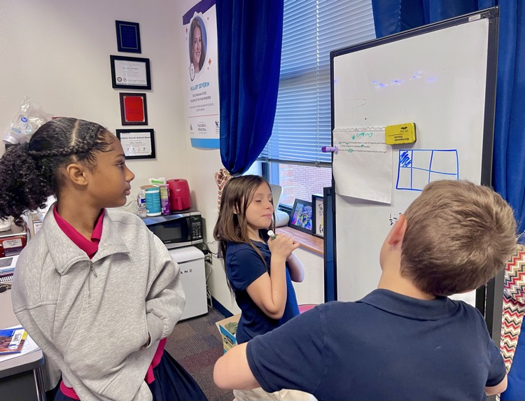 Three students interact with a whiteboard in a classroom. One child holds a marker, another looks on.