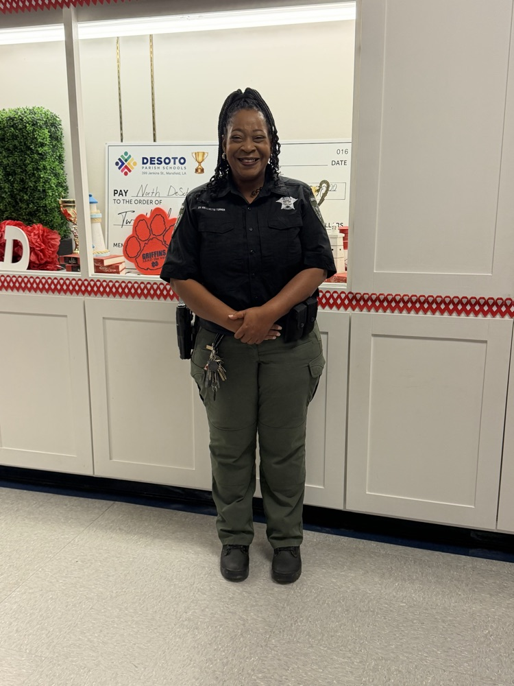 A police officer stands in front of a white counter, hands on hips, wearing a black shirt and green pants.