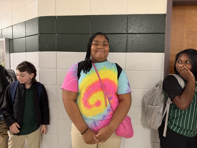 Three students in a hallway, one wearing a tie-dye shirt, another holding a pink bag, and a third with a backpack.
