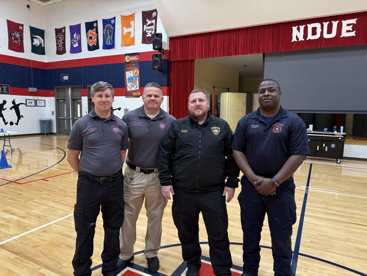 Four men in uniform standing in a gymnasium. The basketball court is visible in the background.