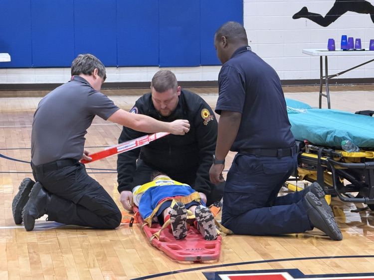 Three men kneeling on a basketball court, one lying on a stretcher, another holding a stick, and a third holding the stretcher.