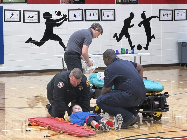 Four adults, one kneeling and three standing, attend to a child lying on a red stretcher on a basketball court.