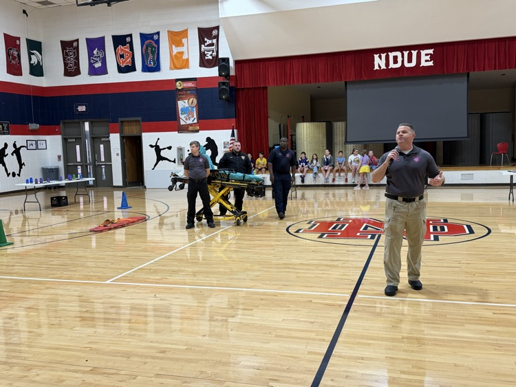 Indoor basketball court with a person speaking into a microphone, two people pushing a stretcher, and two others standing near a table.