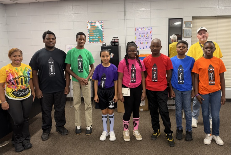 Students in colorful shirts and socks stand in a group in a classroom, next to a man.
