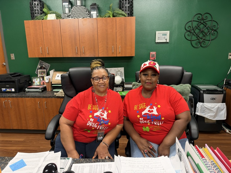Two women in red shirts sit at a desk with folders and papers, with a green wall and cabinets in background.