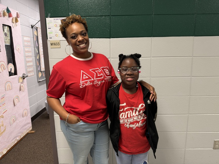 Two people in red t-shirts stand side by side in front of a white and green wall.