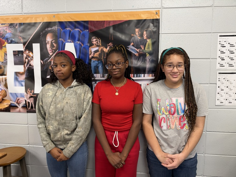 Three students standing close together against a white wall with a banner behind them.
