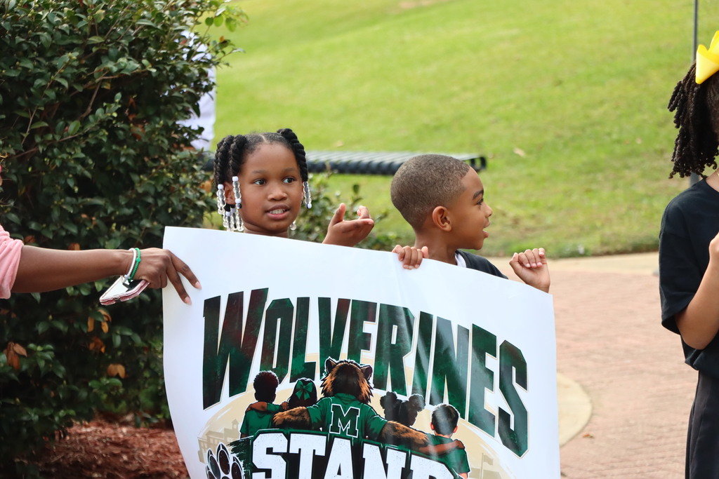 Children holding up a banner that says "WOLVERINES STAND" with an image of people in sports uniforms.