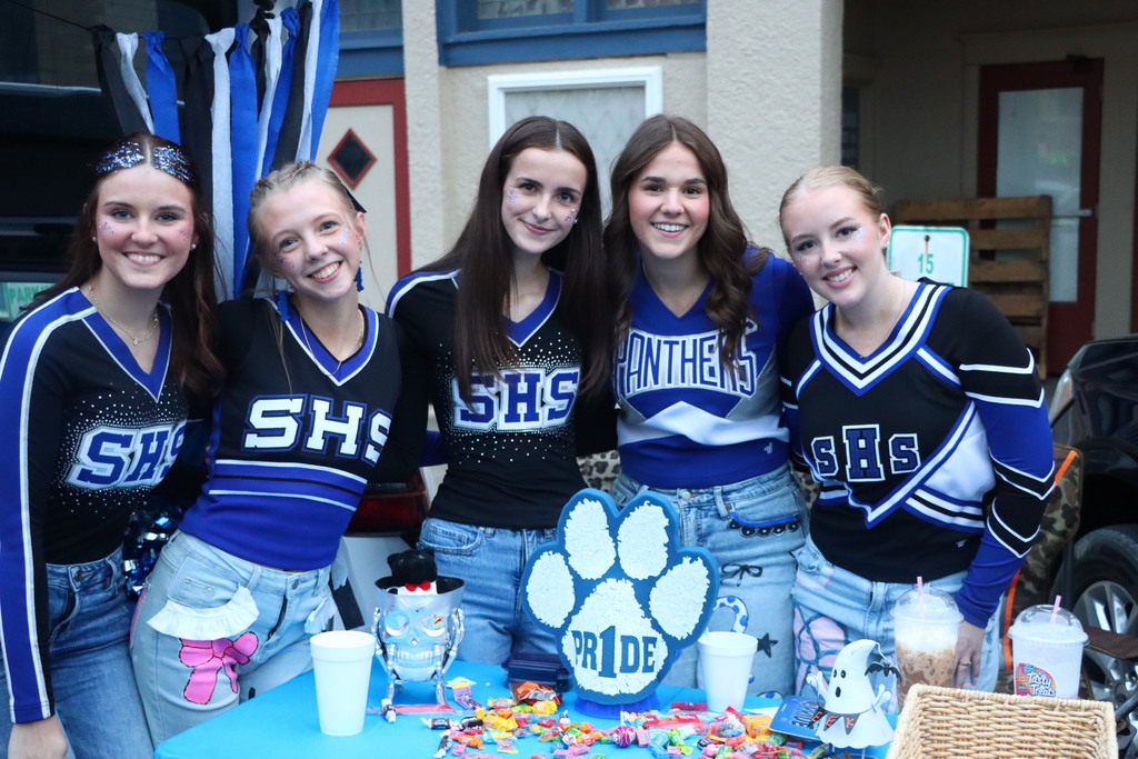 Five cheerleaders stand together outside. Four wear blue and black uniforms with their school's initials. A paw print cake sits on a table with drinks and candy.