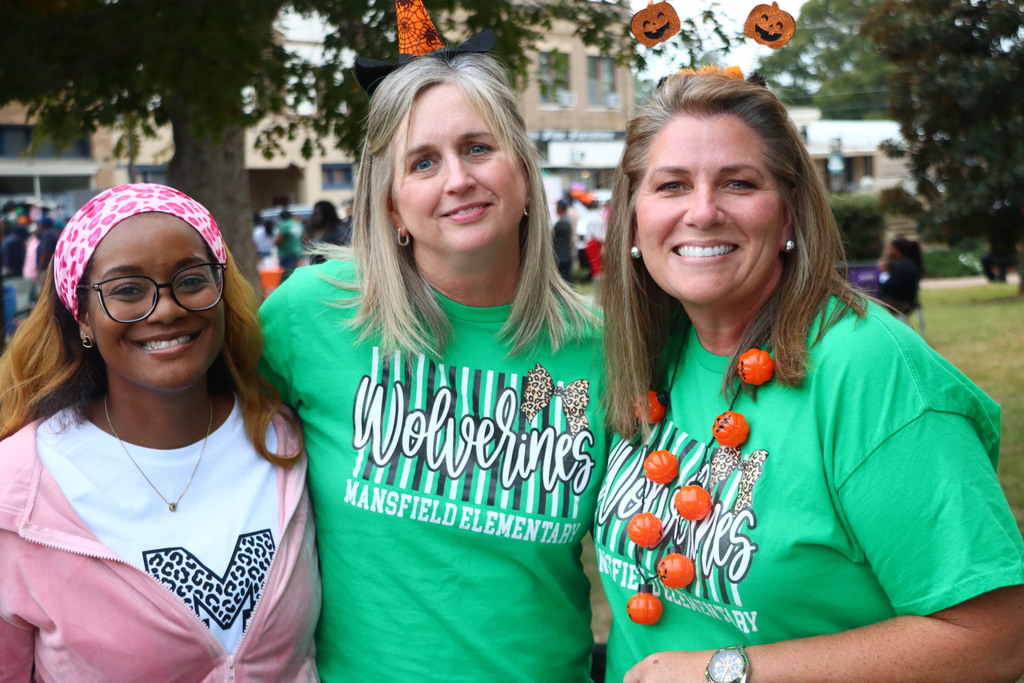 Three smiling adults in green shirts standing under a tree with pumpkins in the background.