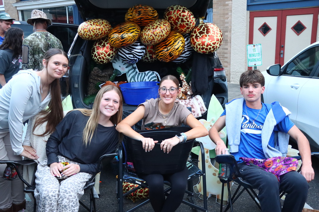 Three girls and one boy sit on chairs next to an open car trunk loaded with large animal-shaped decorations.