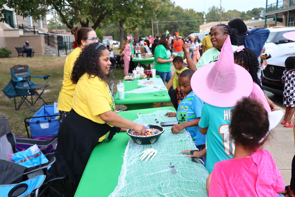 Woman in yellow shirt prepares bowl of candy on green table at school event. Children nearby.