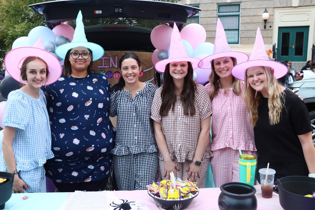 Five women wearing pink hats pose together at a table. Behind them, a car with an open trunk displays balloons.