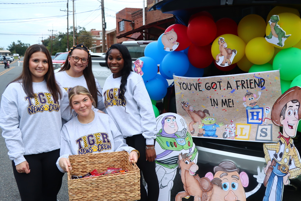 Five girls stand in front of a decorated car. One holds a basket. Balloons and cardboard cutouts are behind them.