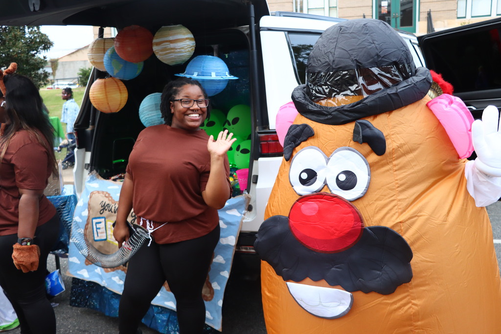 Woman standing near a decorated van, waving, with another person in a Mr. Potato Head costume.