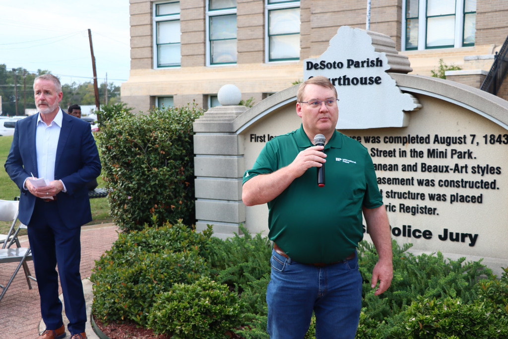 Two men outside a building with a sign; one speaks into a microphone while the other holds a paper.