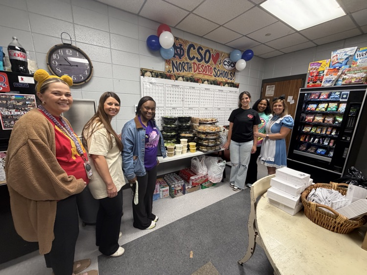 Five adults in costumes stand in a room with tables and a snack machine. One holds a basket of food.