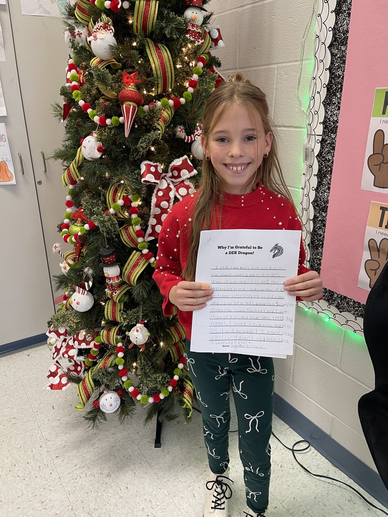 Female student in front of a decorated christmas tree, wearing a red shirt, black pants and holding a writing prompt