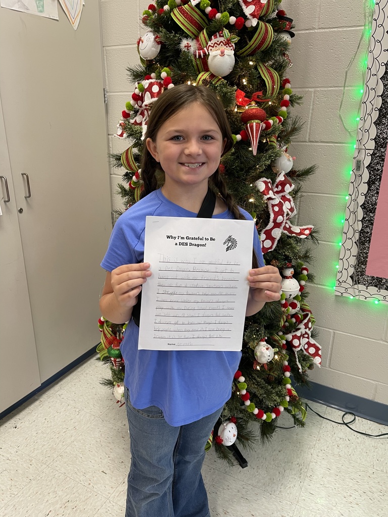 Female student in front of a decorated christmas tree, wearing a blue shirt, denim pants and holding a writing prompt