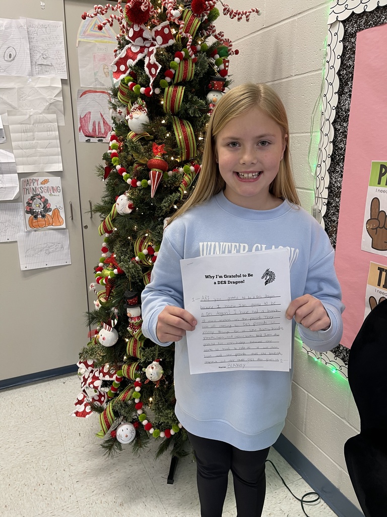Female student in front of a decorated christmas tree, wearing a rlong sleeve ice blue shirt, black pants and holding a writing prompt