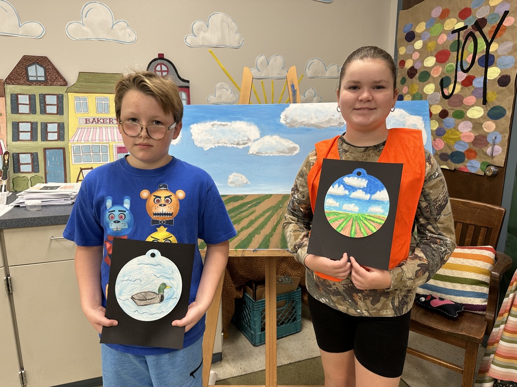 a male des student with a blue shirt holding a picture of a christmas ornament with a duck, a female des student with a camo shirt and an orange vest holding a picture of a christmas ornament depicting a soy bean field