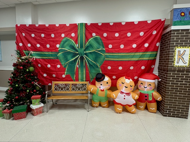 Christmas photo backdrop with a large present wall scene, a Christmas tree with presents on the left, a bench and three blow up gingerbread people