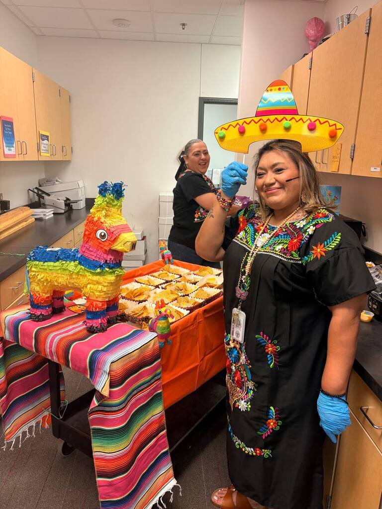 Woman in traditional dress, sombrero, and gloves standing by table with food and pinata.
