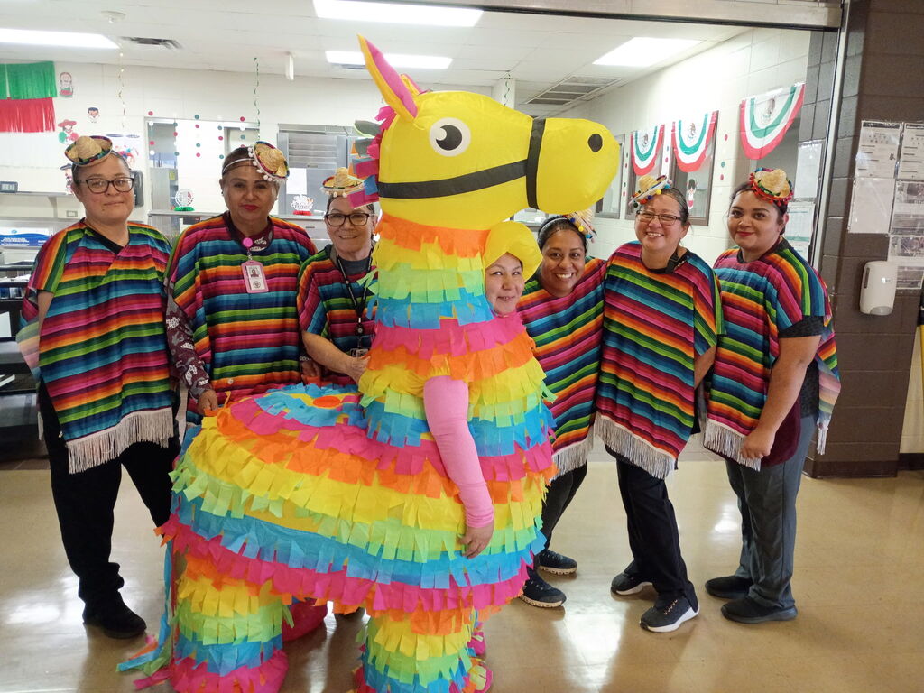 A group of people in multicolored ponchos stands in a room. One person wears a piʇata costume.