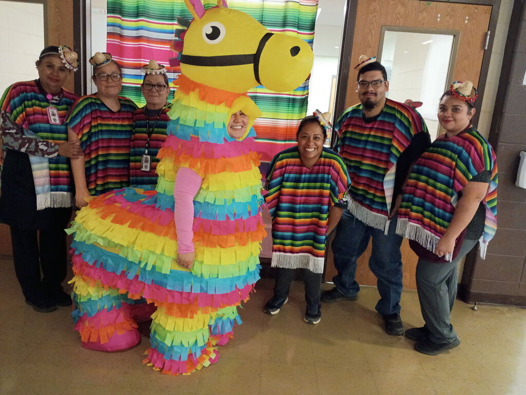 A group of six people dressed in multicolored ponchos pose behind a large, colorful piñata. A door with a mirror is visible in the background.