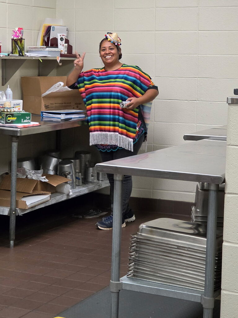 A woman in a colorful poncho stands in a kitchen, gesturing with her hand. Behind her, a table holds items, and a shelf is stacked with boxes.