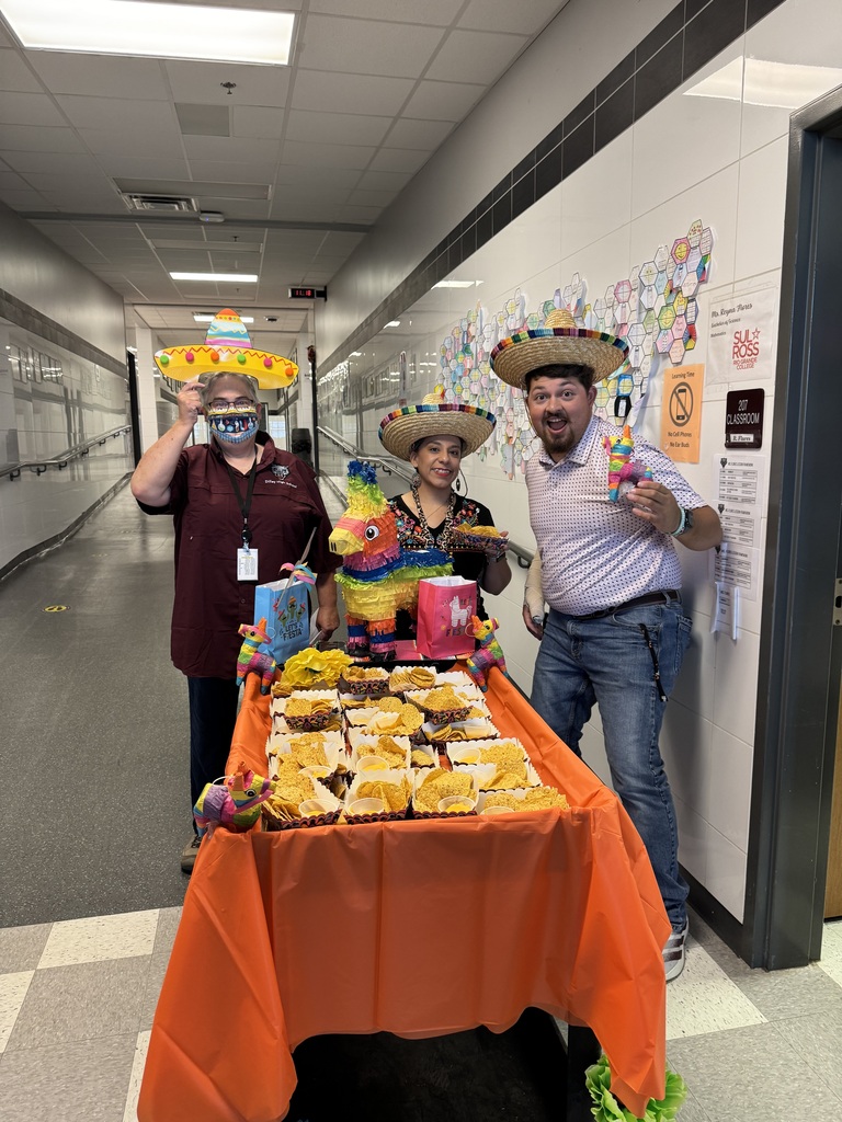 Three people wearing sombreros and masks stand behind a table with food in a hallway.
