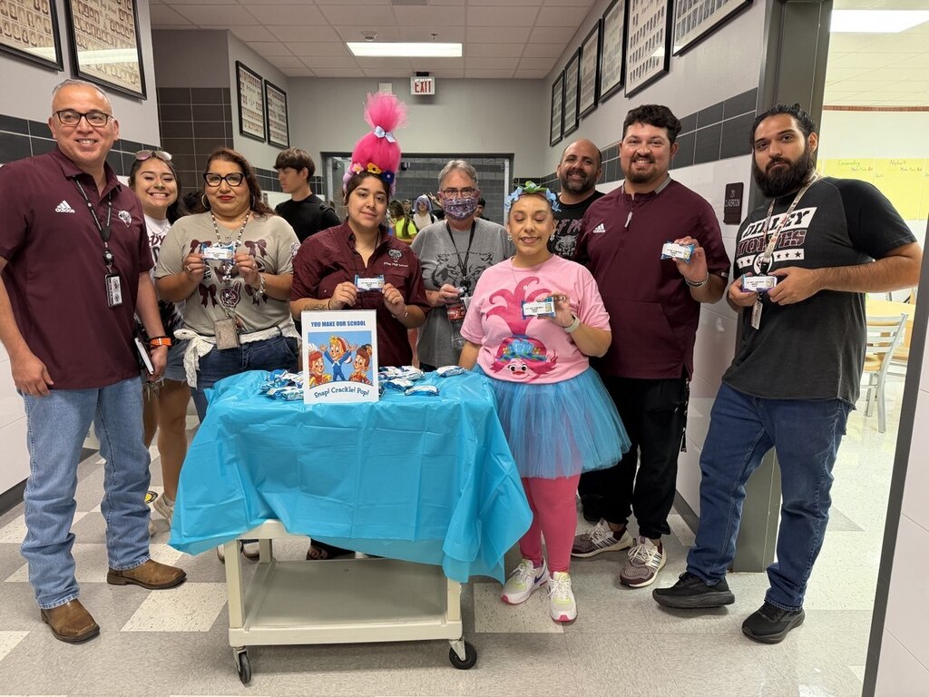 A group of people standing around a table with a blue cover. One person in a tutu holds a book.