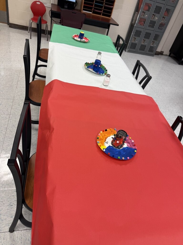 A long table with a red tablecloth. Green and white tablecloth sections, a colorful plate, and a glass. Chairs around the table.