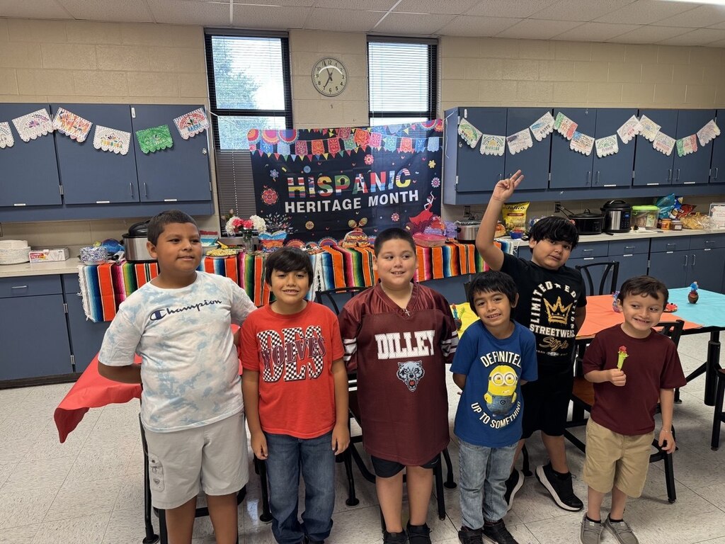 Six children standing in a classroom, with a Hispanic Heritage Month banner behind them and a table in the background.
