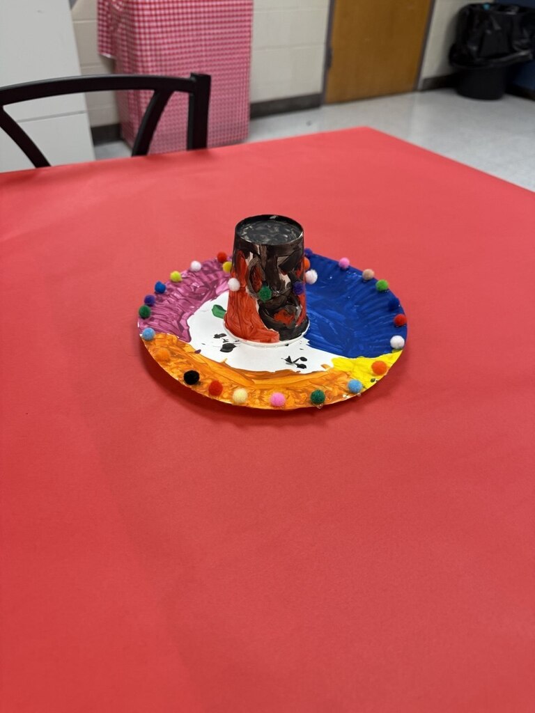 A red tablecloth covers a table, displaying a multicolored paper plate with a cylindrical art piece in the center.