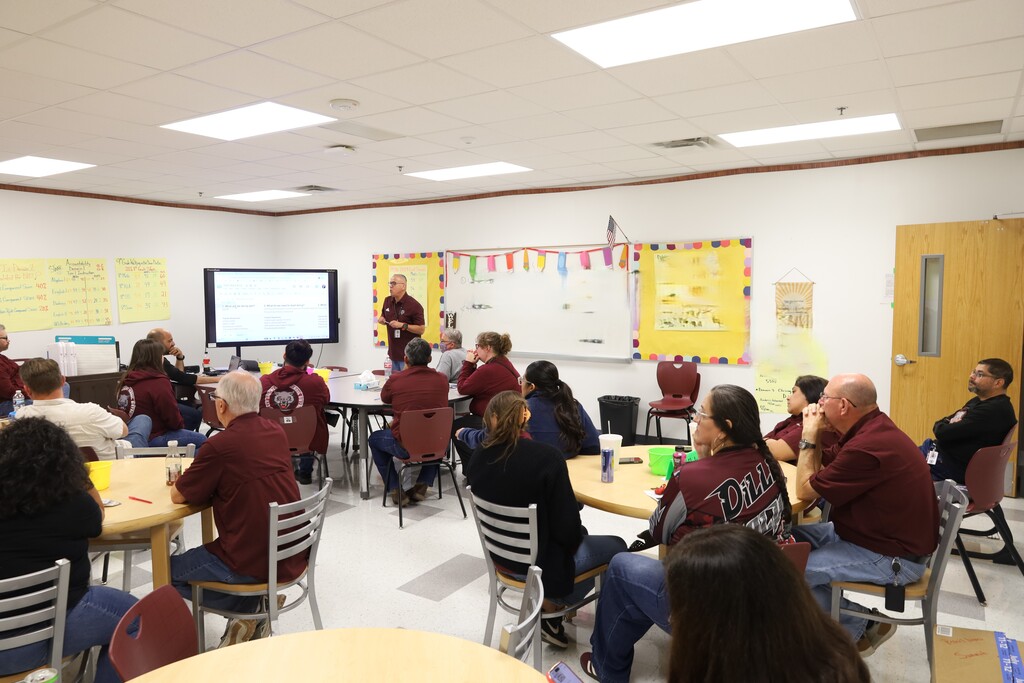A classroom with round tables, people seated, and a person standing before a screen on a wall.