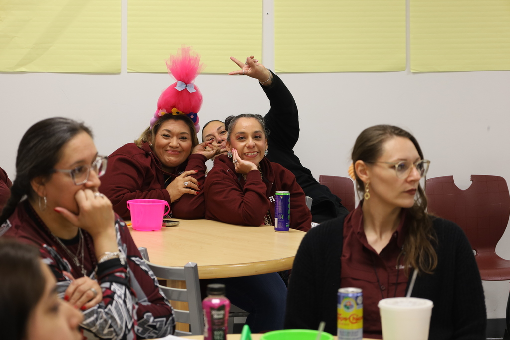 Group of people seated at a table with yellow and red walls. Two people wear pink hair accessories.