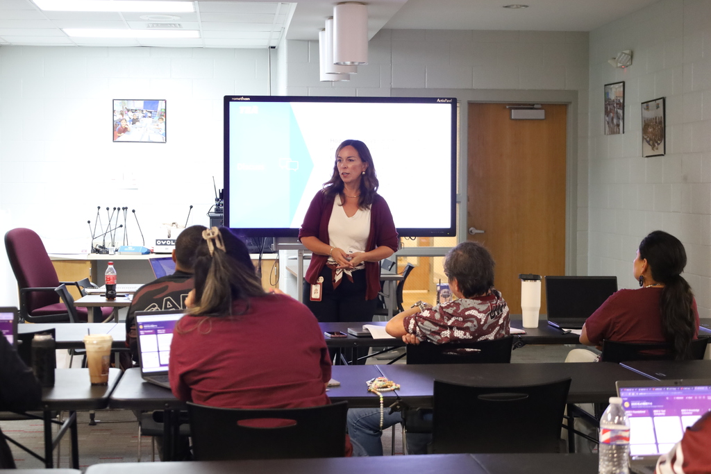 A woman in a white shirt stands in front of a screen, teaching a class of people seated at tables.