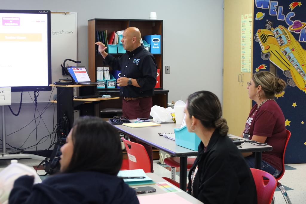 A person stands at a podium, presenting in a classroom. Behind them is a screen and a bookshelf. Three people listen, seated around a desk.