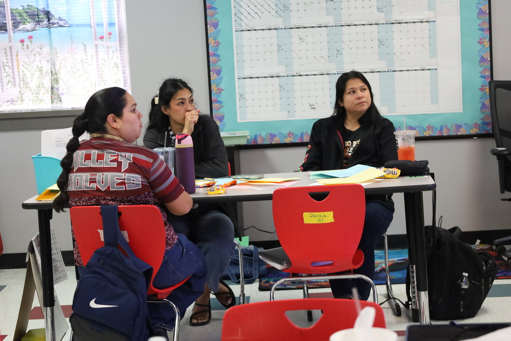 Three people sit at a table with papers and backpacks; a wall with a calendar is behind them.