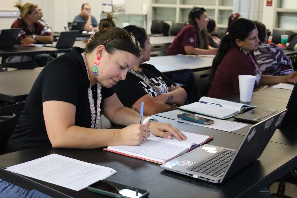 A woman writes in a notebook while seated at a desk, surrounded by others in a classroom.