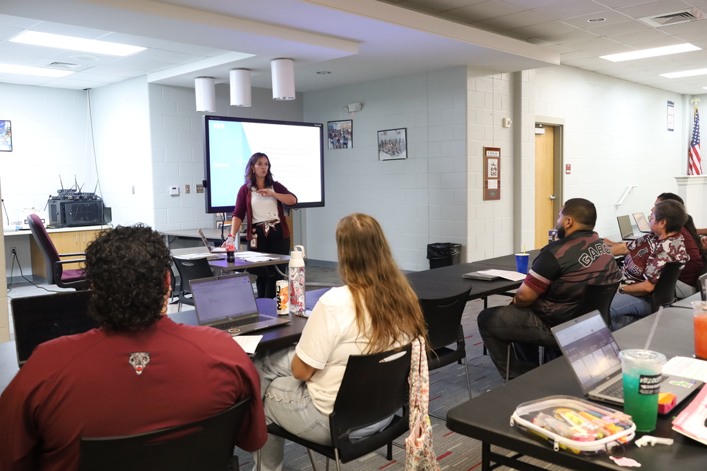 A group of individuals seated around a table in a classroom, listening to a woman speaking in front of a projector screen.