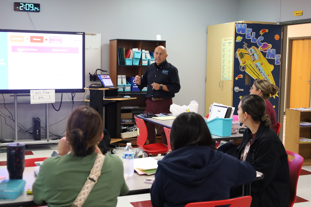 A man presents to a group in a classroom. A screen displays information behind him. People listen.