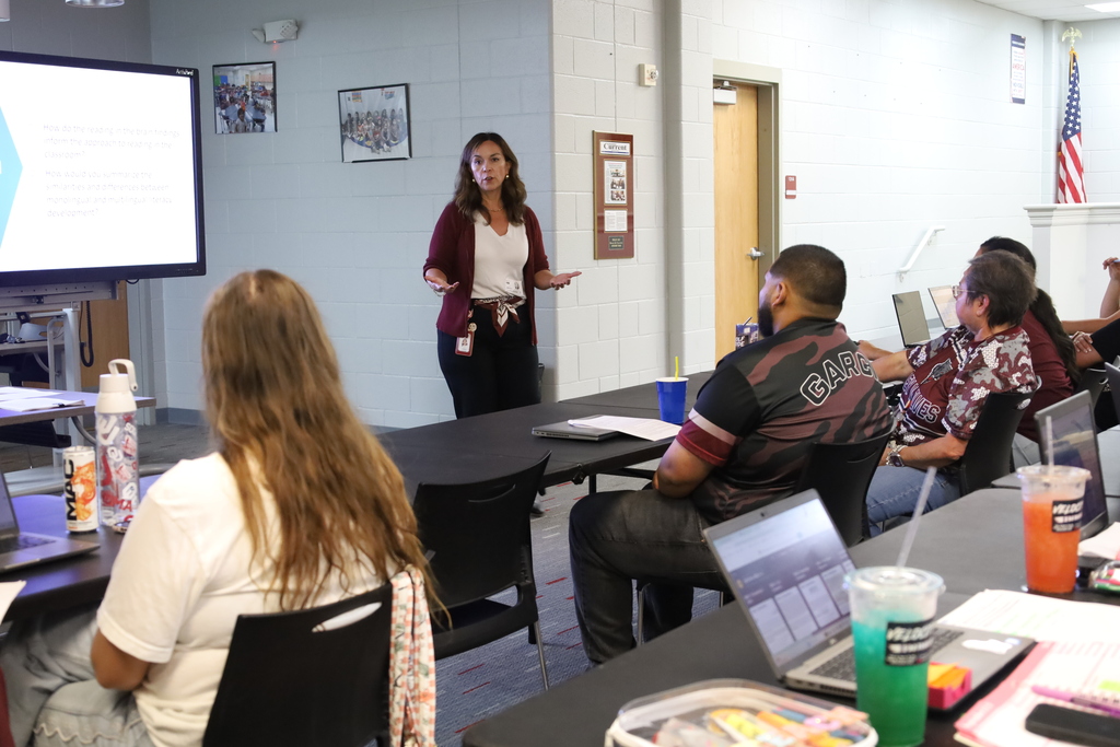 A woman speaks to a small group in a classroom, with attendees seated at tables, laptops open.