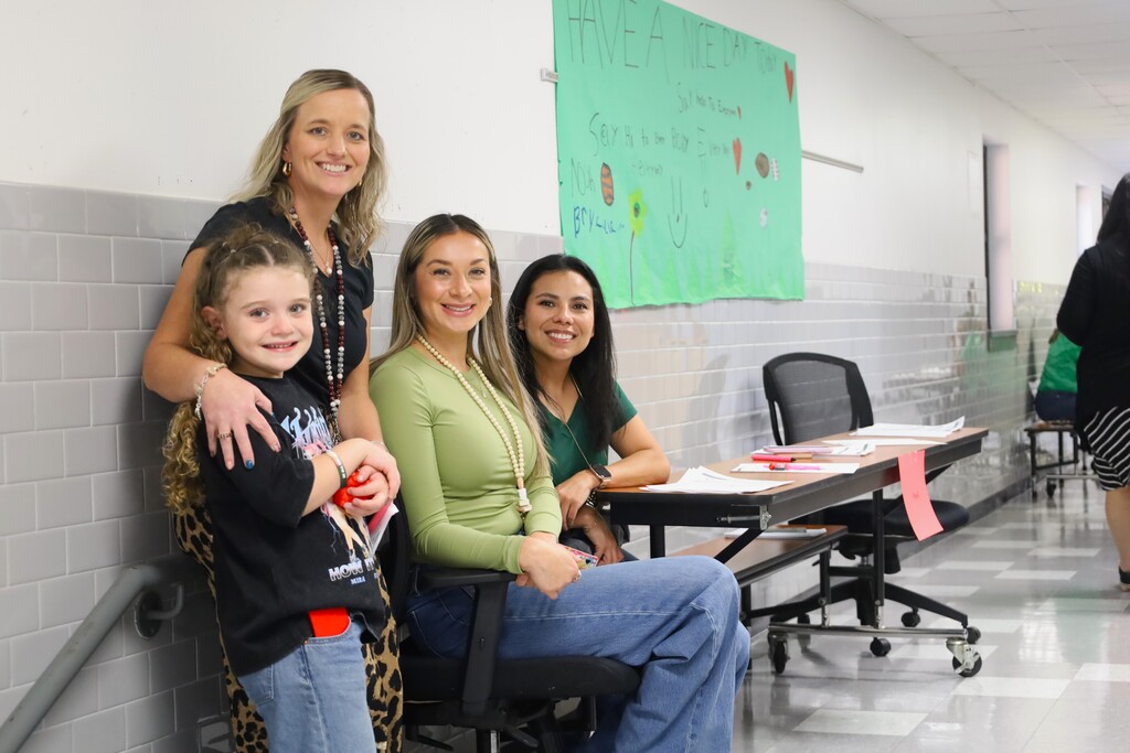 Four women in a hallway. Two seated in chairs, one standing and one child standing behind. Behind them is a table.